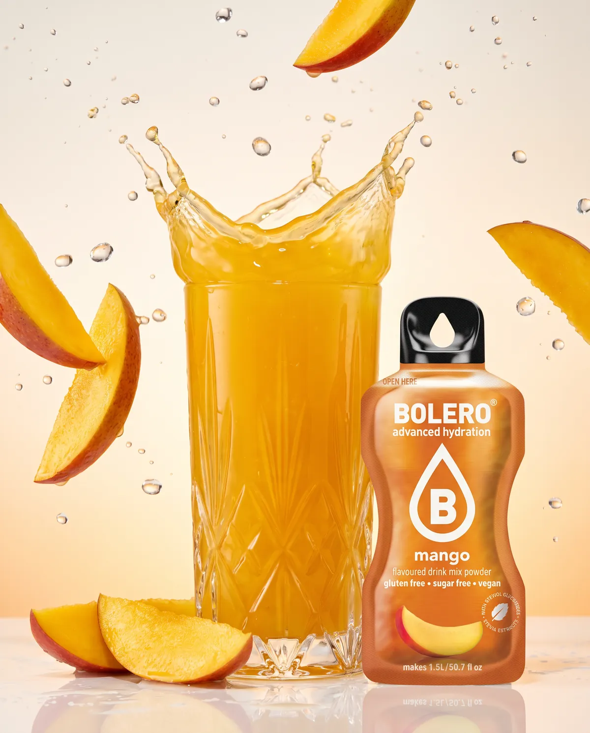 Glas mit goldgelbem Mango-Drink im Splash, fliegende Mangospalten, daneben das Bolero-Päckchen Mango